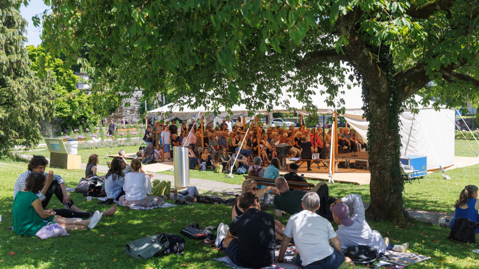 Zuhörendes Publikum sitzt in einem Zelt in einem grünen Park, davor sitzten Menschen im Gras.
